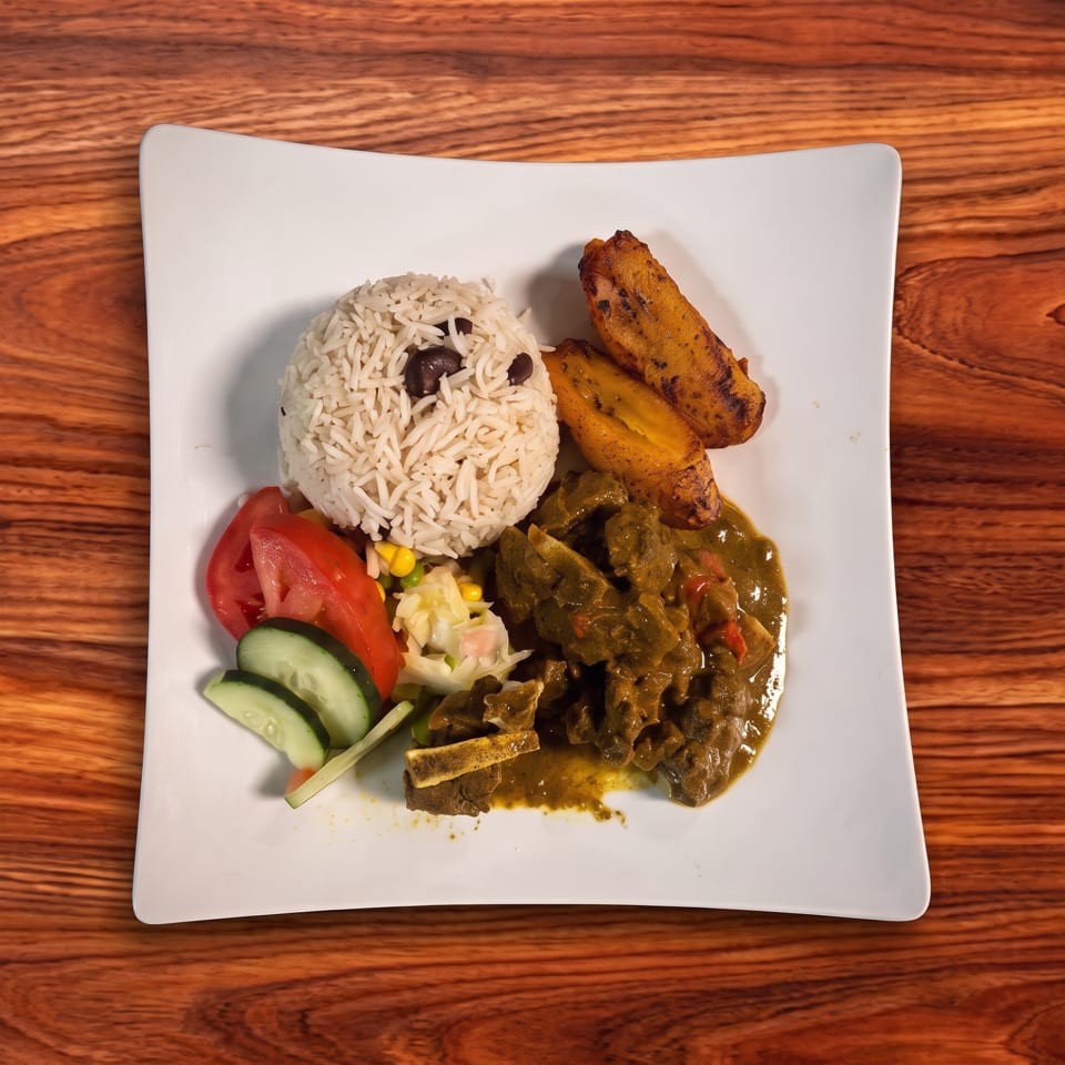 Curried goat with rice and peas, fried plantains, and a side salad on a white plate.