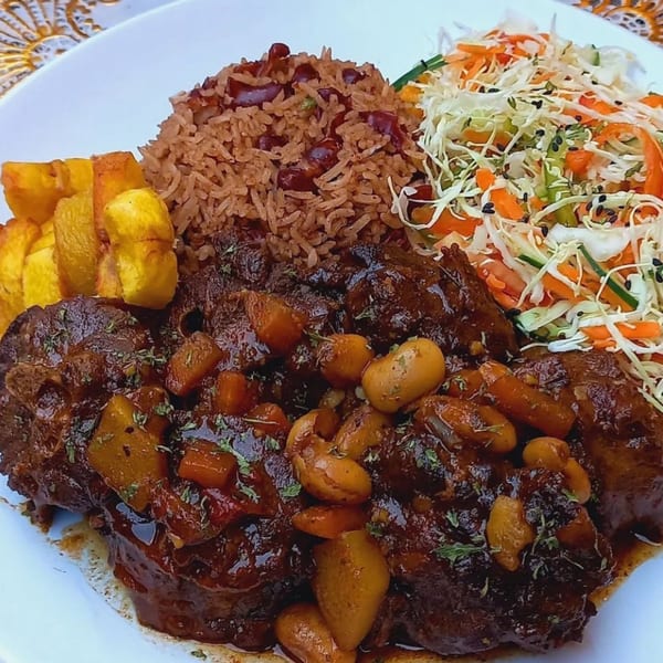 A plate of oxtail stew with rice and peas, fried plantains, and a side salad.