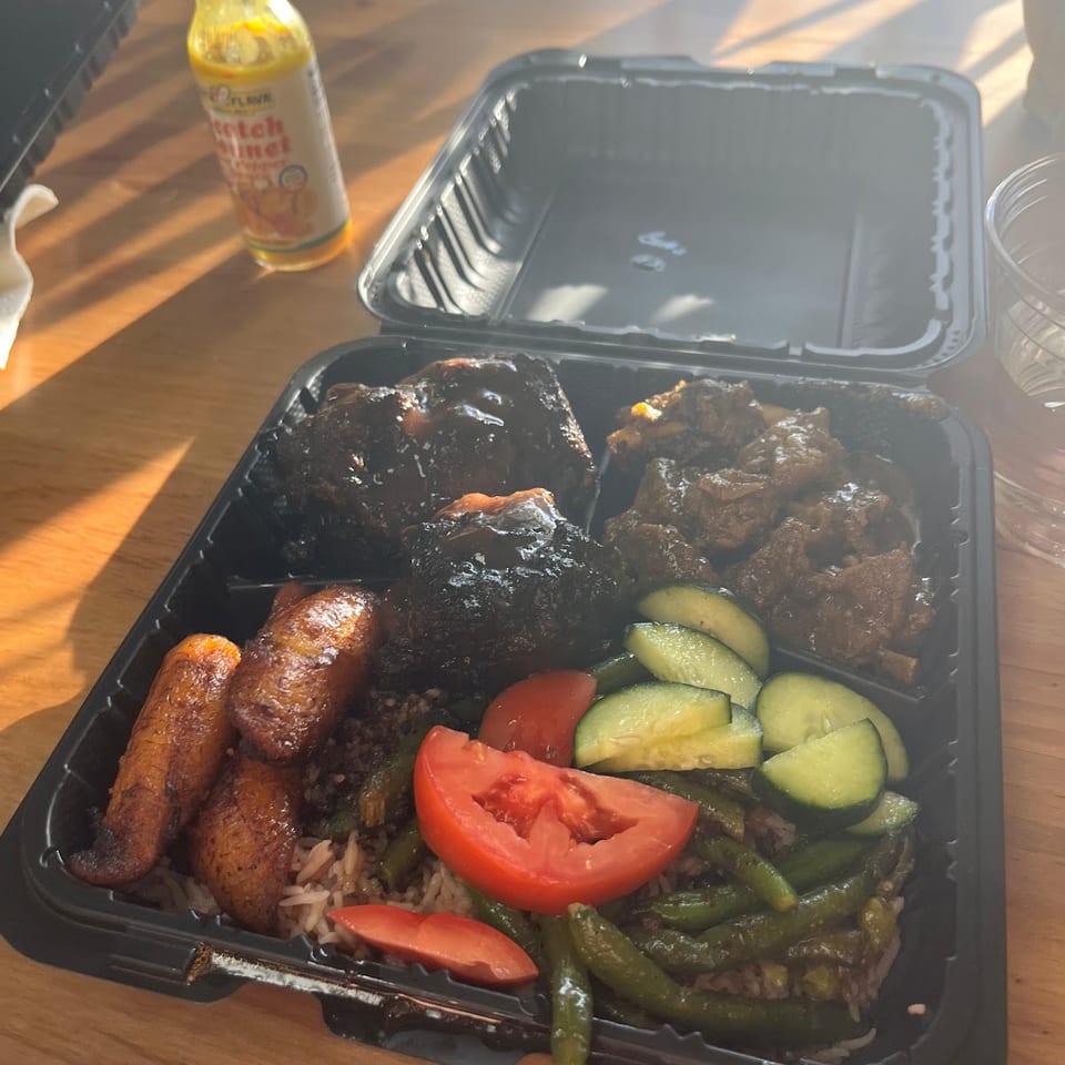 A takeout container filled with jerk chicken, stewed beef, plantains, rice, green beans, tomatoes, and cucumbers.