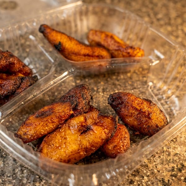 Clear takeout container holding golden fried plantain slices and darker fried pieces on a granite countertop.